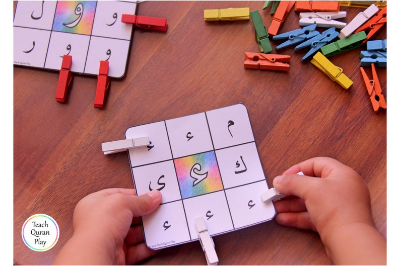 Op de afbeelding: Een witte kaart met gedrukte Arabische letters. De kaart is verdeeld in negen vierkanten. Het middelste vierkant heeft een regenboogkleurig ontwerp. Een kinderhand houdt een witte wasknijper vast. De kaart ligt op een houten tafel met andere wasknijpers die eromheen liggen.