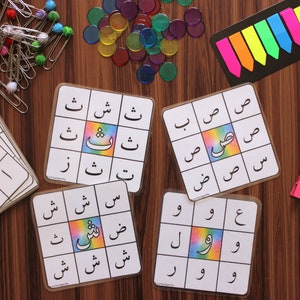 Op de afbeelding: Vier kleurrijke spelborden met Arabische letters en een regenboogachtergrond. De borden zijn omringd door diverse kleurrijke benodigdheden, waaronder clips, tellers en plakbriefjes.