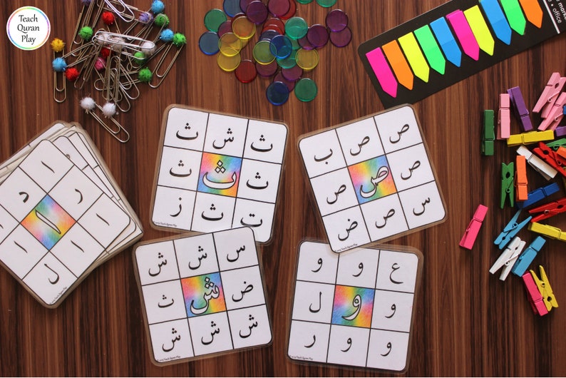 Op de afbeelding: Vier kleurrijke spelborden met Arabische letters en een regenboogachtergrond. De borden zijn omringd door diverse kleurrijke benodigdheden, waaronder clips, tellers en plakbriefjes.