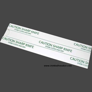 May include: A white rectangular label with the text "CAUTION SHARP KNIFE STORE CLEAN AND DRY" printed in green. The label is angled, and the website address www.USAknifemaker.com is visible at the bottom.