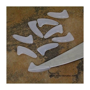 May include: A close-up of a group of clear plastic knife handle scales. The scales are shaped like a crescent moon and are arranged on a brown surface. A silver knife blade is visible in the background.