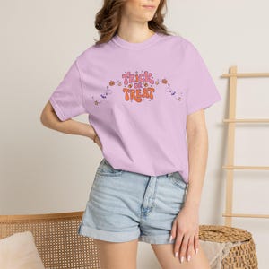 以下が含まれることがあります： ライトパープルの半袖Tシャツ。胸元にはレトロなオレンジ色のフォントで「TRICK OR TREAT」の文字がプリントされています。デザインには、オレンジ色のカボチャ、紫色のコウモリ、黄色の星といった小さなグラフィック要素が曲線状に配置されています。