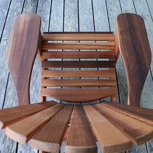 May include: A wooden chair with a slatted back and seat, and curved armrests. The chair is made of natural wood with a reddish-brown color.