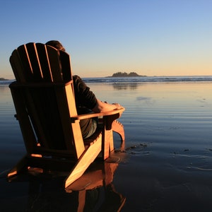 May include: A person sits in a wooden adirondack chair on a sandy beach, facing the ocean. The water is calm and reflects the setting sun.