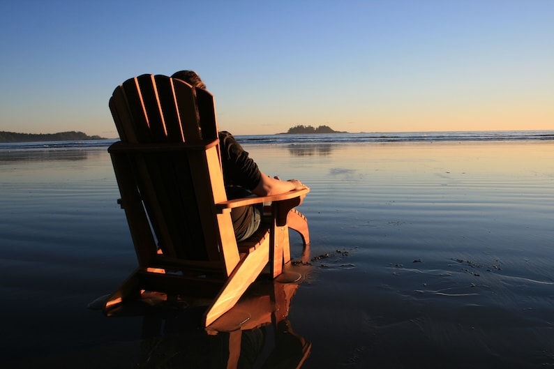 May include: A person sits in a wooden adirondack chair on a sandy beach, facing the ocean. The water is calm and reflects the setting sun.