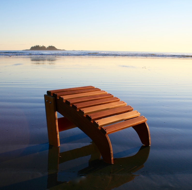 May include: A wooden slatted footrest or stool with a curved design. The footrest is on a sandy beach with the ocean in the background.