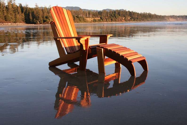 May include: A wooden adirondack chair and footrest on a sandy beach with a reflection in the water.