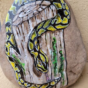 Könnte beinhalten: Handbemalter Stein mit der Darstellung einer grün-gelben Schlange, die sich um einen Baumstamm windet. Die Schlange hat eine gespaltene Zunge und blickt direkt auf den Betrachter.