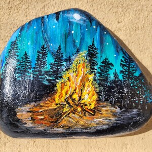 Könnte beinhalten: Handbemalter Stein mit einer Darstellung einer Feuerstelle mit einem leuchtend orangefarbenen und gelben Feuer in der Mitte, umgeben von einem dunklen Wald mit einem blauen und weißen Sternenhimmel darüber.