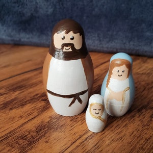 May include: A set of three wooden nesting dolls painted to resemble the Holy Family. The largest doll is brown and depicts Joseph, the middle doll is blue and depicts Mary, and the smallest doll is white and depicts baby Jesus.