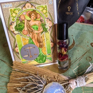 May include: A tarot card depicting the Libra zodiac sign with a woman holding a scale. The card is surrounded by flowers and stars. A small bottle of essential oil with dried flowers inside is next to the card. A broom with a crescent moon charm is in the foreground.