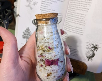 May include: A glass bottle with a cork stopper filled with white bath salts, dried herbs, and pink flower petals. The bottle is labeled "PIMPERNEL" and "Pimpinella anagalis, P. sp." The text is from a book on magical herbs.