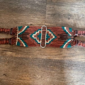 Puede incluir: Un cinch de caballo trenzado marrón, verde y blanco con hebillas plateadas. El cinch está hecho de cuerda trenzada y tiene un patrón geométrico.