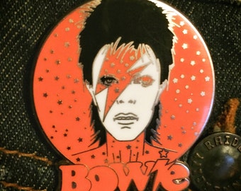 Puede incluir: Un pin de esmalte rojo y blanco con un retrato de David Bowie con el texto "BOWIE" en letras naranjas. El fondo es un círculo rojo con estrellas blancas.