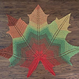 May include: A handmade, open-weave shawl in the shape of a maple leaf. The shawl features a gradient of colors, transitioning from red at the base to green, yellow, and finally, brown at the tips. The shawl is made of yarn.
