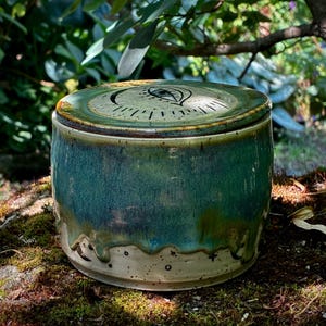 May include: A cylindrical ceramic jar with a lid featuring a hand-drawn eye design. The upper portion has a glossy teal and green glaze that drips down over a speckled cream-colored base. The base is decorated with small black celestial symbols.