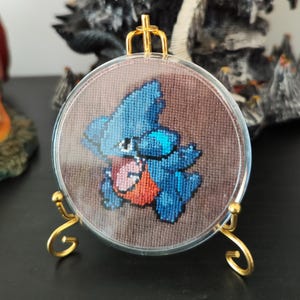 May include: A round, cross-stitched artwork featuring a blue cartoon character with red and blue accents. The artwork is displayed in a clear, circular frame, held by a gold-colored metal stand. The background is a brown fabric.