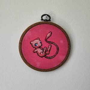 May include: A pink and white embroidered picture of a pink Pokemon character with blue eyes, framed in a brown wooden hoop.