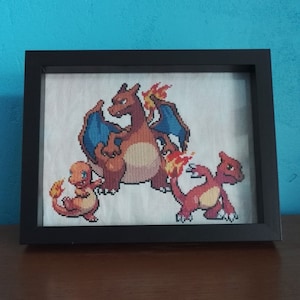 Może przedstawiać: Ramka z haftem krzyżykowym przedstawiająca trzy postacie Pokemonów: Charmandera, Charmeleona i Charizarda. Postacie są przedstawione w swoich charakterystycznych pomarańczowych i czerwonych kolorach, a Charizard ma niebieskie skrzydła.