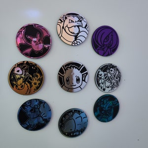 May include: Nine round, silver-coloured Pokémon trading card game tokens with various Pokémon characters. The tokens feature a variety of colours, including purple, blue, gold, and silver.