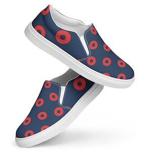 May include: A pair of slip-on shoes featuring a dark blue fabric upper with a repeating pattern of solid red circles. The shoes have white elastic side panels and a thick white rubber sole, providing a clean contrast to the dark blue design.
