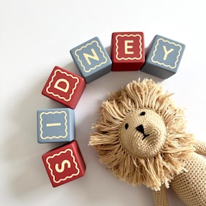 May include: Six wooden blocks arranged in an arc, alternating between red and grey colours, each featuring a gold letter spelling SIDNEY inside a scalloped border. Beside the blocks is a beige crocheted lion toy with a textured fringe mane.