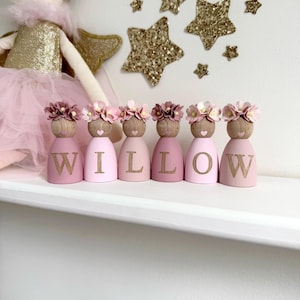 May include: Six wooden peg dolls arranged in a row, each wearing a floral crown and painted in shades of pink. Each doll features a single letter from the name WILLOW in gold, with a small heart painted below the face. A fabric doll is visible.