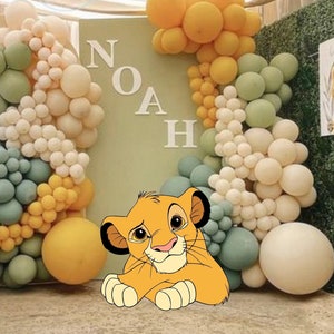 Puede incluir: Un expositor decorativo para eventos con un gran panel verde salvia con las letras blancas NOAH, rodeado por un arco de globos en tonos verde salvia, crema y amarillo. Una estatua de elefante gris se sitúa a la izquierda y un cachorro de león de dibujos animados está en primer plano.