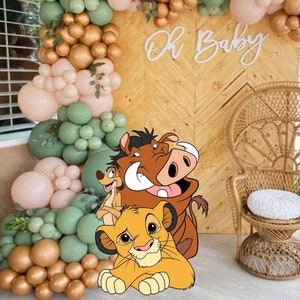 Puede incluir: Un fondo decorativo para eventos con una pared de madera en espiga y un arco de globos en verde salvia, rosa suave y oro metálico. El texto Oh Baby está montado en la pared. Se incluyen una silla de mimbre y un gráfico de Simba, Timon y Pumbaa.