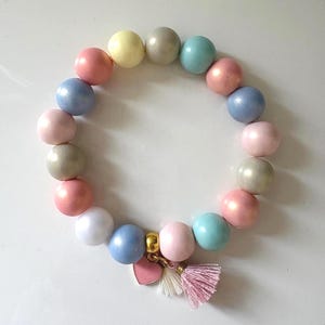 Può includere: Un braccialetto di perline dai colori pastello con perline rotonde rosa, blu, gialle e grigie. Il braccialetto include un ciondolo dorato con un cuore rosa e un nappa rosa.