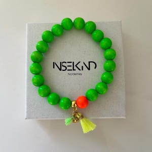 Könnte beinhalten: Ein grünes Perlenarmband mit einer leuchtend orangenen Perle und goldenen Quastenanhängern. Das Armband liegt auf einer weißen Box mit dem Text "INSEKIND Norderney".