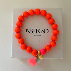 Könnte beinhalten: Ein leuchtend orangefarbenes Perlenarmband mit einem rosa Anhänger und zwei Quasten. Das Armband befindet sich in einer weißen Box mit dem aufgedruckten Text "INSEKIND" und "Norderney".