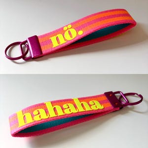 Puede incluir: Dos llaveros con un diseño a rayas rosas, naranjas y turquesas. Uno tiene la palabra "nö." en amarillo, y el otro dice "hahaha" en amarillo. Ambos tienen un cierre y un anillo de metal morado.