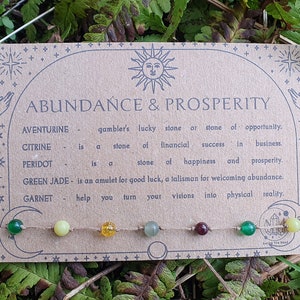 May include: A card with the words "ABUNDANCE & PROSPERITY" and gemstone descriptions. A string of beads in various colours, including green, yellow, and red, is attached. The card is set against a backdrop of green foliage.