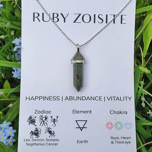 May include: A Ruby Zoisite pendant necklace with a silver chain. The pendant is a green and red crystal. A card behind the necklace reads "RUBY ZOISITE" and "HAPPINESS | ABUNDANCE | VITALITY". Zodiac, Element, and Chakra symbols are also on the card.