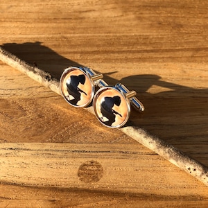 May include: A pair of silver cufflinks with a round, cream-colored background and a black silhouette of a person. The cufflinks are on a wooden surface.
