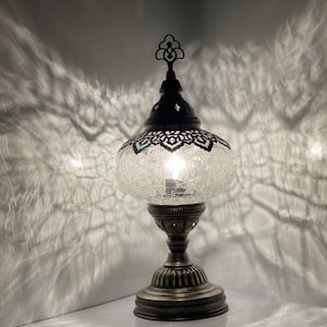 Może przedstawiać: Lampa stołowa z kloszem z przezroczystego szkła w kształcie kuli z pękniętym wykończeniem. Lampa ma metalową podstawę z ozdobnym wzorem i zwieńczeniem na górze.