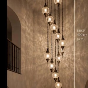 Può includere: Un lampadario a più luci con 15 globi in vetro craquelé sospesi ad altezze variabili da un supporto circolare a soffitto. L'apparecchio ha una caduta totale di 400 cm (4 m). Ogni globo è fissato a una catena e a una struttura in metallo scuro.