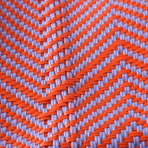 May include: Close-up of a woven plastic mat with a checkered pattern in orange and purple.