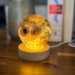 May include: A decorative illuminated sphere with dried flowers and gold flakes encased in clear resin. The sphere sits on a round wooden base, emitting a warm, yellow glow. The lamp is powered by a cord.