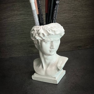 May include: White plaster bust of a man with a short, wavy hairstyle, used as a pencil holder.  The bust is holding a variety of makeup pencils, including black, brown, and white.