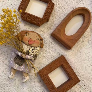 May include: Three wooden picture frames in various shapes, including a square and an oval. A handmade doll with a floral scarf and a bouquet of yellow flowers is also present. The frames are brown and have textured edges.