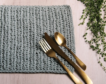 May include: A gray crocheted placemat with a herringbone pattern, featuring a gold fork, knife, and spoon.