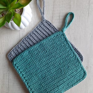 Könnte beinhalten: Zwei handgehäkelte Topflappen. Einer ist türkis, der andere grau. Beide haben eine Schlaufe zum Aufhängen. Die Topflappen sind quadratisch und haben eine gerippte Textur. Die Topflappen sind etwa 20 cm groß.