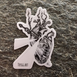 Puede incluir: Pegatina en blanco y negro de un corazón atravesado por una espada, con una cara de calavera y una boca dentada. La pegatina tiene el texto "TRYGG.ART" en la parte inferior.