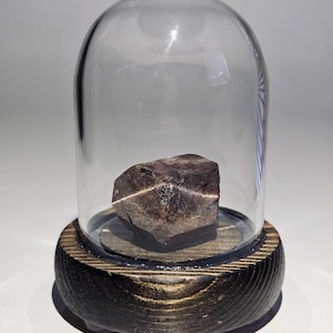 May include: A dark brown, faceted, geometric rock is displayed under a glass dome on a wooden base.