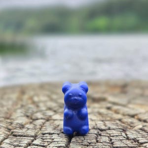 May include: A small, solid blue bear-shaped figurine with rounded ears, a smooth surface, and a defined snout. The bear is standing upright, featuring a simple, molded design with visible facial features including eyes and a nose on its front side.