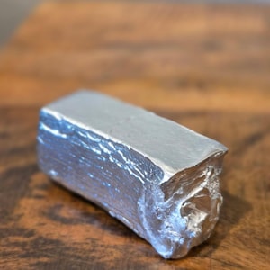 May include: A rectangular silver-colored metal ingot with a rough, textured surface. The object has a solid, block-like form with irregular, jagged edges on one end, showcasing a metallic sheen and a dense, heavy appearance throughout its structure.