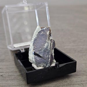 May include: A dark, irregularly shaped mineral specimen, partially coated in a light-colored substance, is displayed in a clear, hinged plastic case. The case sits within a black, rectangular base, suggesting a presentation or display setting.