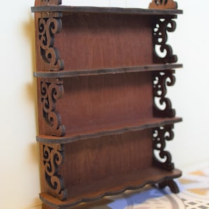 Pu&ograve; includere: Un'unit&agrave; a scaffale in legno con tre ripiani e decorazioni a volute sui lati. L'unit&agrave; a scaffale &egrave; di colore marrone e ha un aspetto rustico.